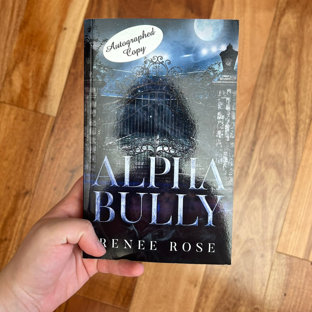 A white person's hand holding a paperback. The book cover features a metal car gate. In the background is the night sky and full moon. There is an "Autographed Copy" sticker on the corner. In blue text is the title "Alpha Bully." On the bottom in white text is "Renee Rose"