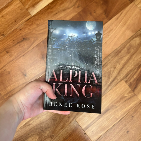A white person's hand holding a paperback. The book cover features a metal car gate. In the background is the night sky and a full moon. In pink text is the title "Alpha King." On the bottom of the cover in white text is "Renee Rose"