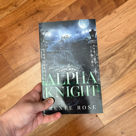 A white person's hand holding a paperback. The book cover features a metal car gate. The night sky and full moon are in the background. In green text is the title "Alpha Knight." In white text on the bottom of the cover is "Renee Rose"