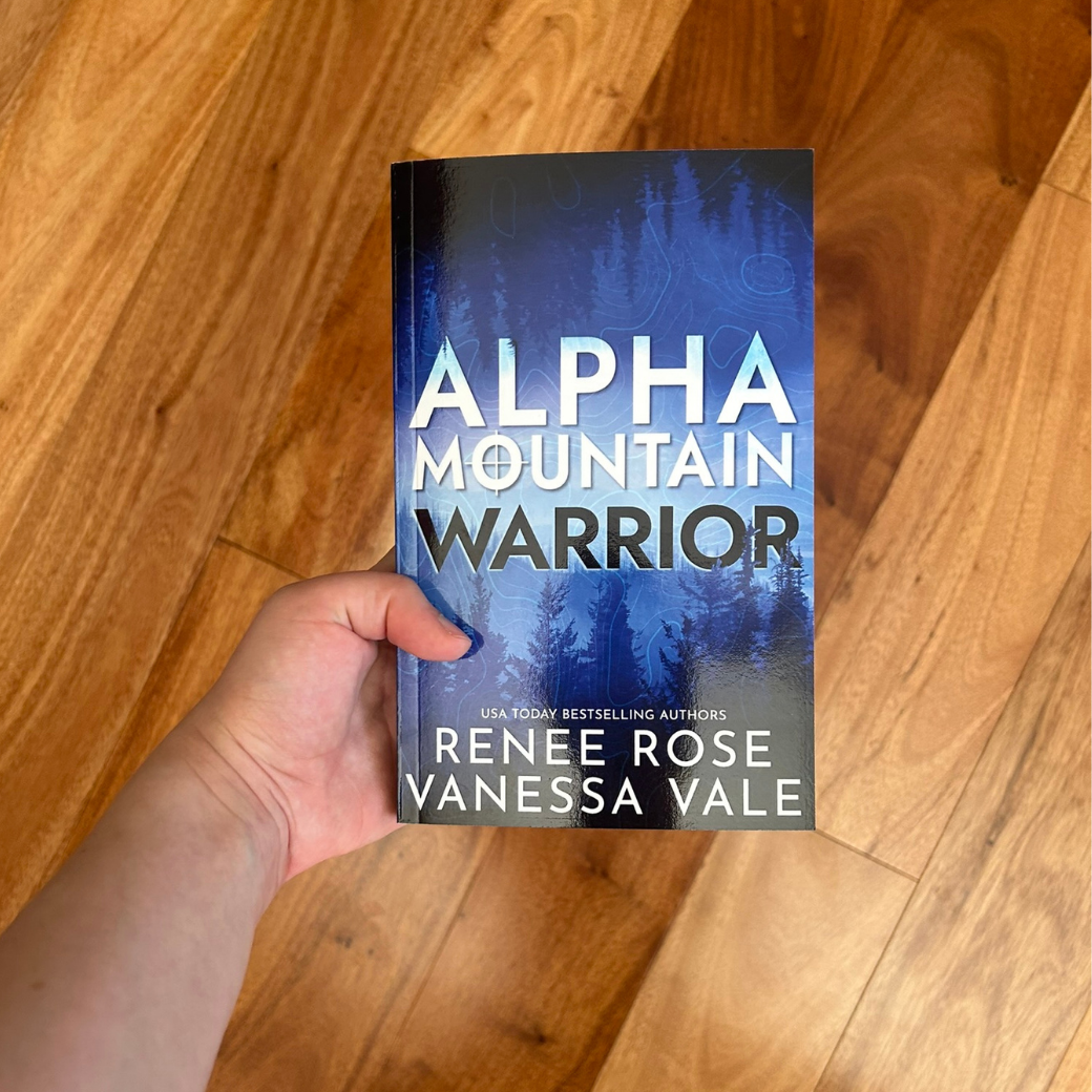 A white person's hand holding a paperback. The book cover is blue and featues a forest. In white and black bold text is the title "Alpha Mountain Warrior." On the bottom of the cover in white text is "USA Today Bestselling Authors -- Renee Rose Vanessa Vale"