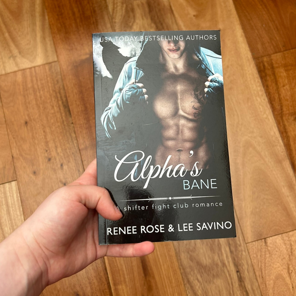 A white person's hand holding a paperback. The cover features a buff, white, shirtless, tattooed man in a black cloak. In the background is a full moon and outline of a wolf. On the top of the cover in white text is "USA Today Bestselling Authors." In white text is the title "Alpha's Bane A shifter fight club romance." On the bottom in white text is "Renee Rose & Lee Savino"