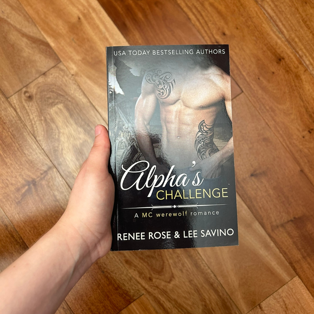 A white person's hand holding a paperback. The book cover features a shirtless, buff, tattooed white man. In the background is the outline of a wolf. On the top of the cover in white text "USA Today Bestselling Authors." In white and yellow text is the title "Alpha's Challenge A MC werewolf romance." On the bottom in white text is "Renee Rose & Lee Savino"