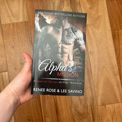 A white person's hand holding a paperback. The book cover features a white shirtless, buff man with a shoulder tattoo. In the background is fire and the outline of a wolf. On the top of the cover in white text is "USA Today Bestselling Authors." In white and red text is the title "Alpha's Mission A special forces shifter romance." On the bottom in white text "Renee Rose & Lee Savino"