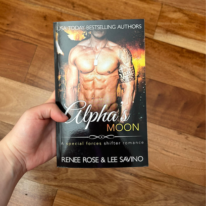 A white person's hand holding a paperback. The book cover features a buff, shirtless, white man with a shoulder tattoo and wearing a dog tag. In the background is fire and the outline of a wolf. On the top of the cover in white text is "USA Today Bestselling Authors." In white and gold text is the title "Alpha's Moon A special forces shifter romance." On the bottom of the cover in white text is "Renee Rose & Lee Savino"