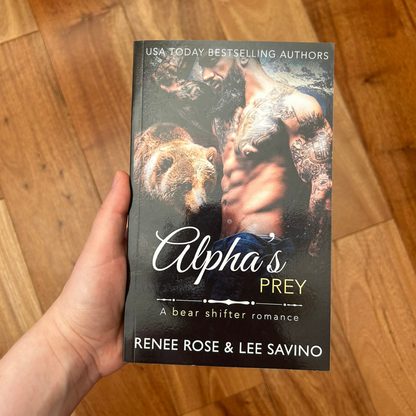 A white person's hand holding a paperback. The cover features a buff, shirtless, tattooed man with a black beard. Behind him is a brown bear. On the top of the cover in white text is "USA Today Bestselling Authors." In white and yellow text is the title "Alpha's Prey A bear shifter romance." On the bottom of the cover in white text is "Renee Rose & Lee Savino"