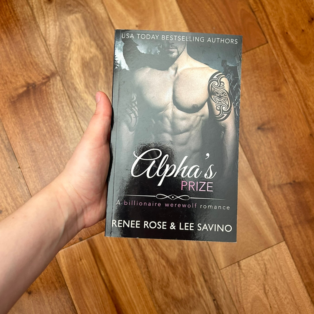 A white person's hand holding a paperback. The book cover features a buff, shirtless, white man. He has arm tattoos. In the background is a full moon and the outline of a wolf. On the top of the cover in white text is "USA Today Bestselling Authors" In white and pink text is the title "Alpha's Prize A billionaire werewolf romance." On the bottom in white text is "Renee Rose & Lee Savino"