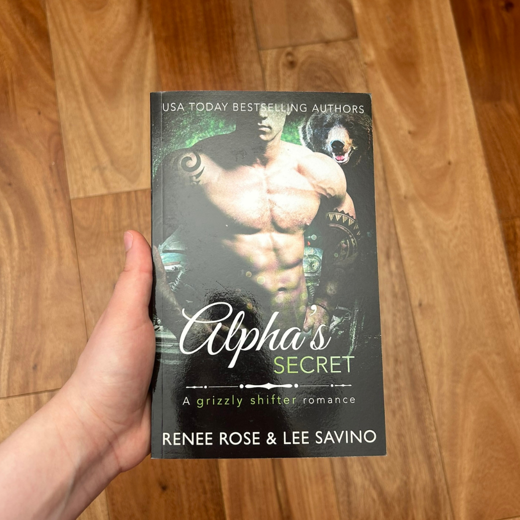 Hand holding a paperback book. The cover features a shirtless, buf, tattooed white man. Behind him is a bear. On the top of the cover in white text is "USA today Bestselling Author." In white and green text is the title "Alpha's Secret A Grizzly Shifter Romance." On the bottom of the cover in white text is "Renee Rose & Lee Savino"