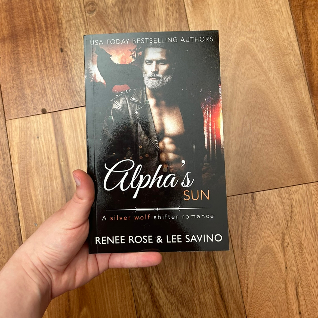 A white person's hand holding a paperback. The book cover features a shirtless, buff man in a leather hacket. He has grey hair and a bushy grey beard. In the background is the outline of a wolf. On the top of the cover in white text is "USA Today Bestselling Authors." In white and orange text is the title "Alpha's Sun A silver wolf shifter romance." On the bottom in white text "Renee Rose & Lee Savino"