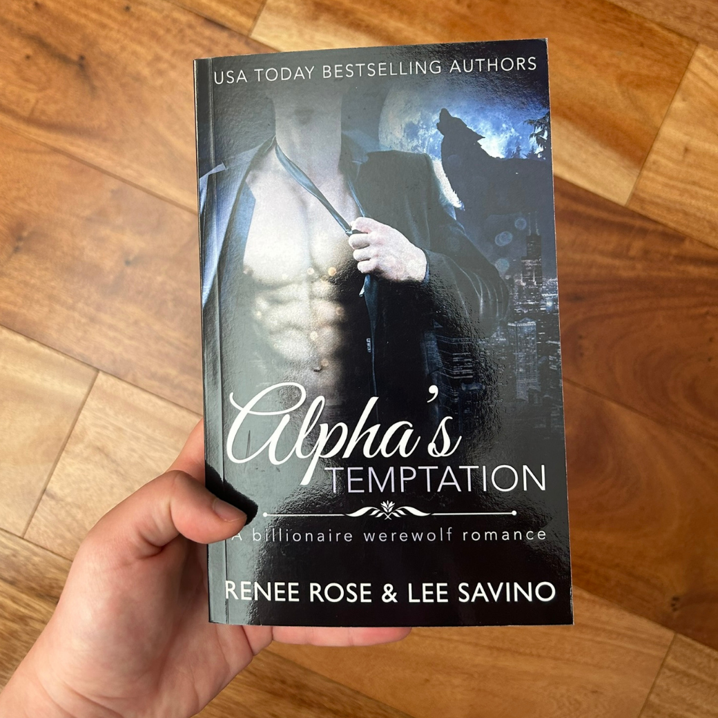 A white person's hand holding a paperback. The cover features a buff shirtless whtie man wearing a black suit jacket. He is loosening a black tie. In the background is a full moon and the outline of a wolf. In white text on the top of the cover is "USA Today Bestselling Authors." In white text is the title "Alpha's Temptation A billionaire werewolf romance." On the bottom of the cover in white text is "Renee Rose & Lee Savino"