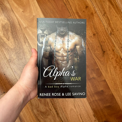A white person's hand holding a paperback. The book coer features a buff, shirtless, tattooed man with a chain hanging on his neck. In teh background is a tiger. On the top of the cover in white text is "USA Today Bestselling Authors." In white and yellow text is the title "Alpha's War A bad boy Alpha romance." On the bottom in white text is "Renee Rose & Lee Savino"
