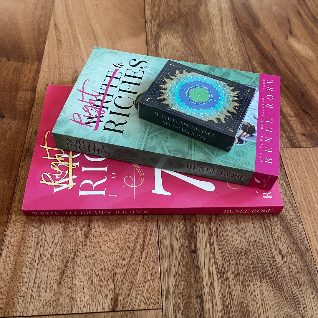 Two author abundance books by Renee Rose stacked on a wooden floor: 'Write to Riches' in mint green with pink script lettering, and 'The  Write to Riches Journal' in bright pink with white lettering, with an Author Affirmation card box featuring a blue and gold circular design resting on top.