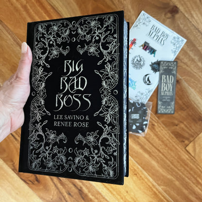Book titled 'Big Bad Boss' by Lee Savino & Renee Rose held by a hand on a wooden surface with other items.