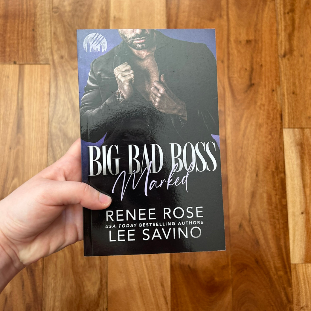 A white person's hand holding a paperback. The book cover's background is indigo. It features a shirtless man who is wearing a black suit jacket. In white and lavender text is the title "Big Bad Boss Marked." On the bottom of the cover in white text is "Renee Rose -- USA Today Bestselling Authors -- Lee Savino"