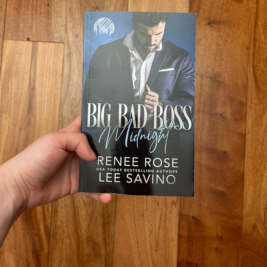 A white person's hand holding a paperback. The book cover has a blue background and features a man in a white button down and blue suit. He is adjusting his cuff. In white and blue text is the title "Big Bad Boss Midnight." On the bottom of the cover in white text is "Renee Rose -- USA Today Bestselling Authors -- Lee Savino"