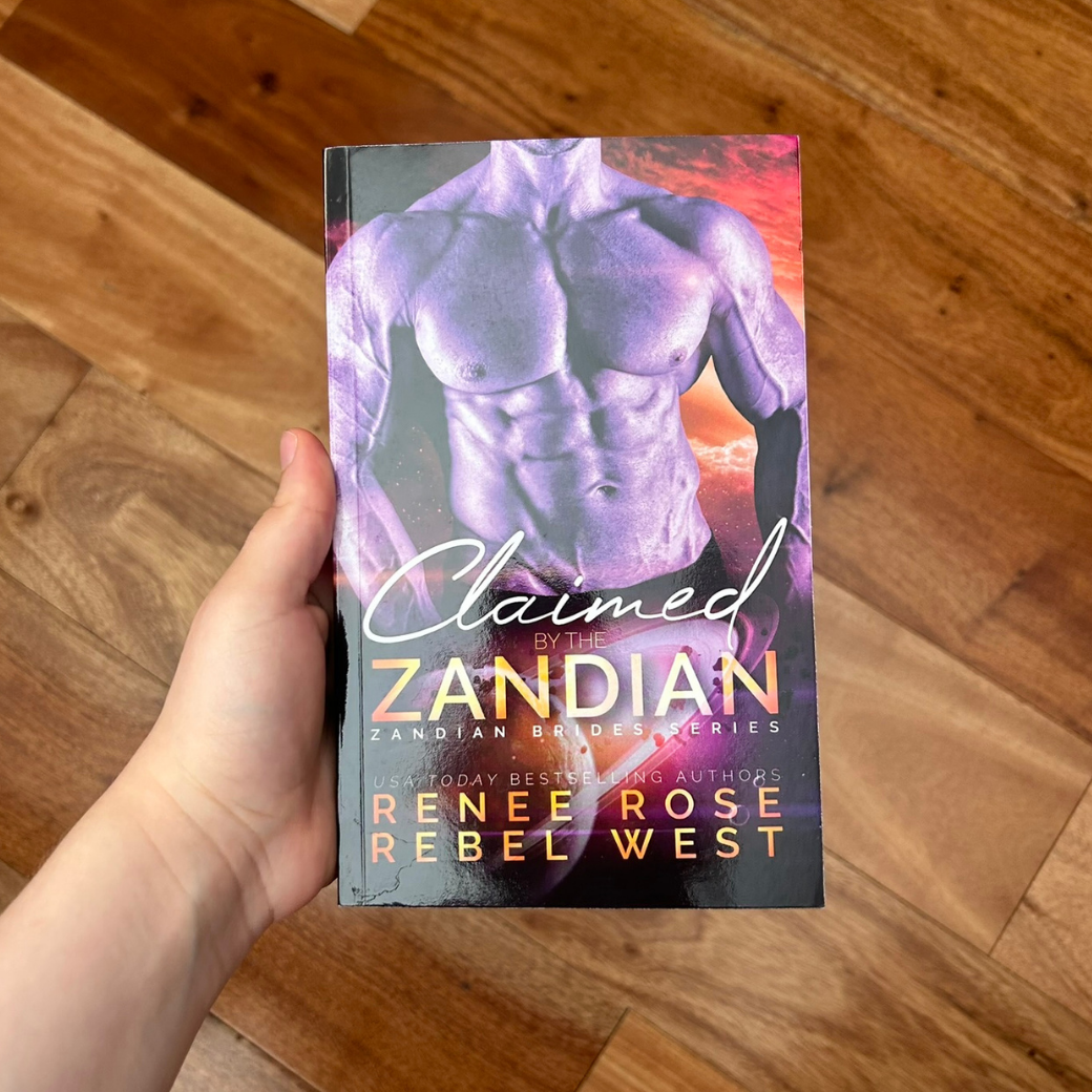 A white person's hand holding a paperback. The book cover features a buff, shirtless white man in purple lighting. In the background is a sunset and a ringed planet. In white and gold text is the title "Claimed by the Zandian Zandian Brides Series." On the bottom in white and gold text is "USA Today Bestselling Authors -- Renee Rose Rebel West"