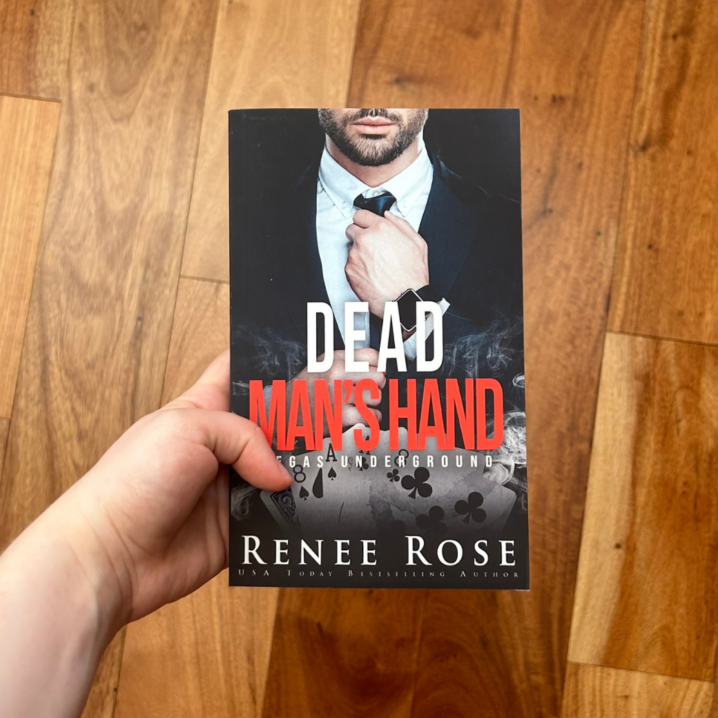 A white person's hand holding a paperback. The cover features a man with scruff in a black and white suit. He is adjusting his tie. In white and red text is the title "Dead Man's Hand Vegas Underground." On the bottom of the cover in white text is "Renee Rose -- USA Today Bestselling Author"