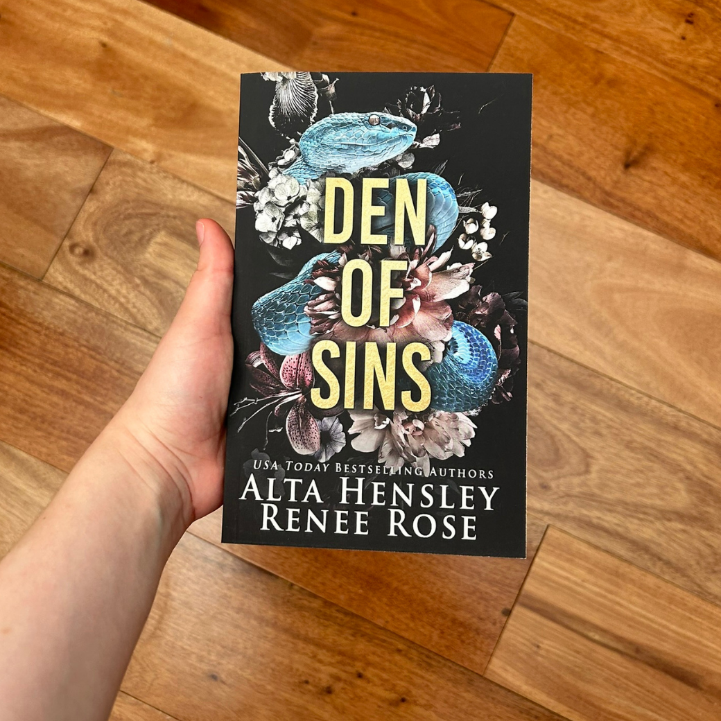 A white person holding a paperback. The book cover has a black background with a blue snack and flowers in stark contrast. On top of the snake in gold text is the title "Den of Sins." On the bottom of the cover in white text is "USA Today Bestselling Authors -- Alta Hensley Renee Rose"