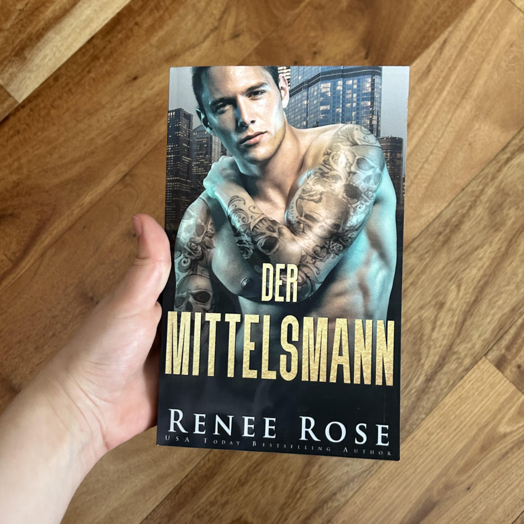 A white person's hand holding a paperback. The book cover features a buff, shirtless, tattooed white man. Behind him is a city. In gold text is the title "Der Mittelsmann." On the bottom of the cover in white text is "Renee Rose -- USA Today Bestselling Author"