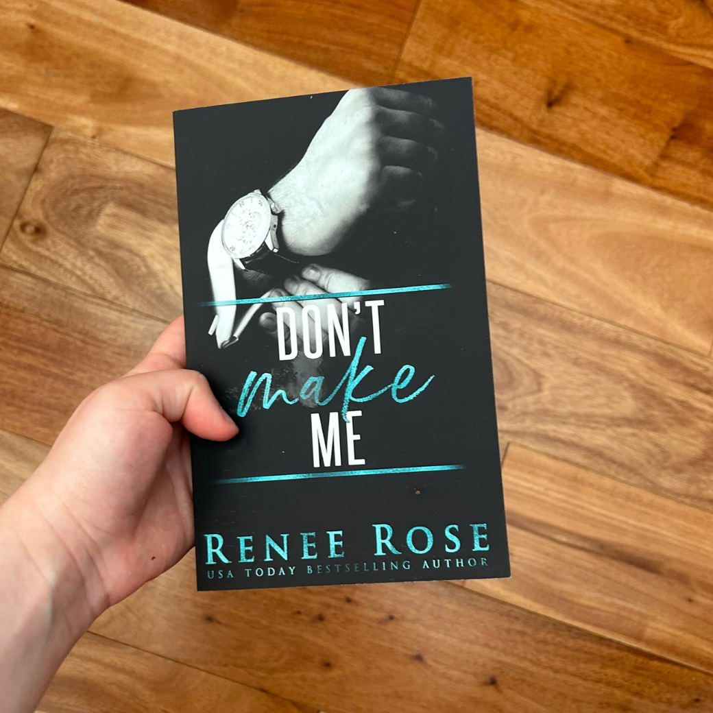 A white person's hand holding a paperback. The book cover has a black background and features a suited man's hands clasping a watch. In white and blue text is the title "Don't Make Me." On the bottom of the cover in blue text is "Renee Rose -- USA Today Bestselling Author"