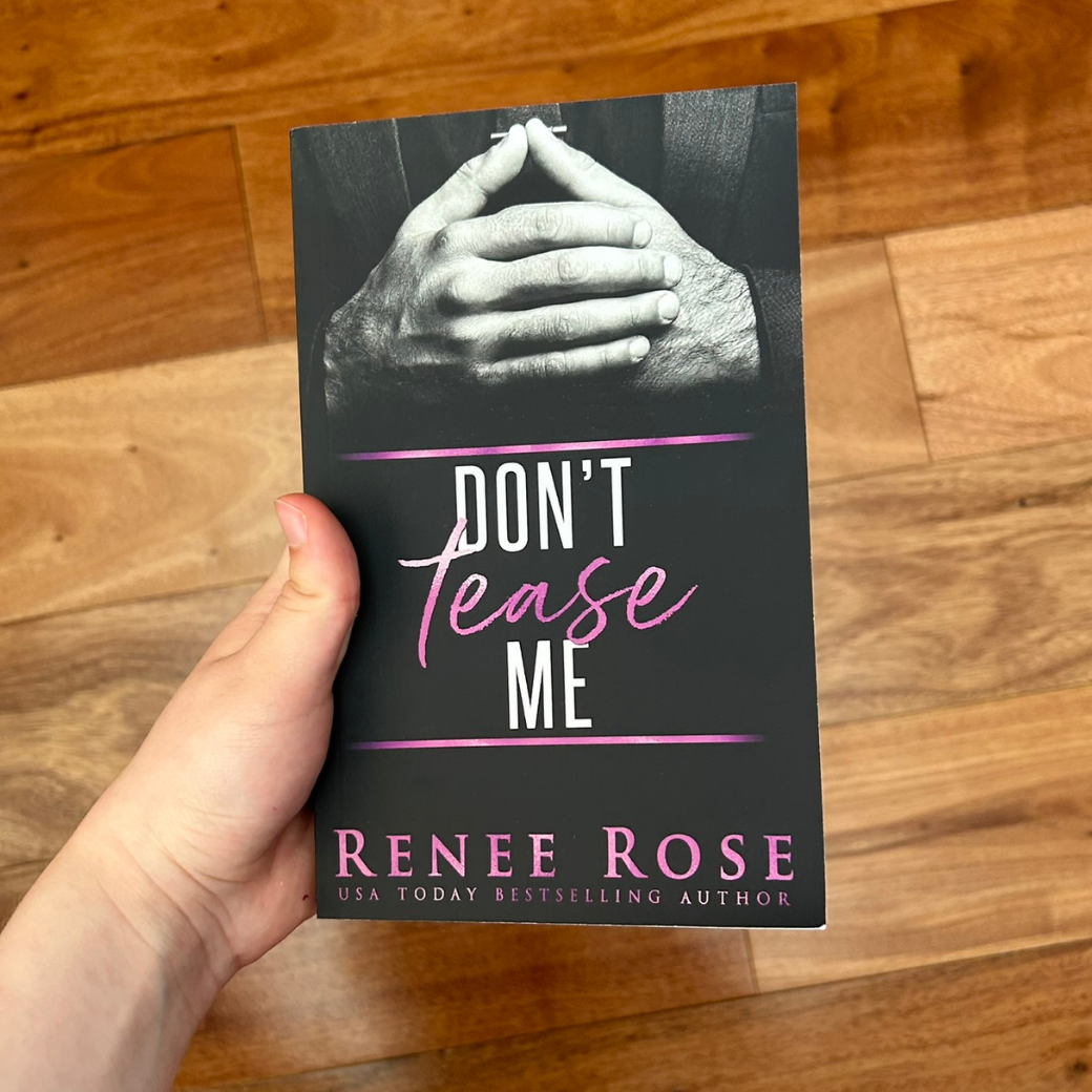 A white person's hand holding a paperback. The book cover is black and features a black suited man's hands clasped in front of himself. In white and pink text is the title "Don't Tease Me." In the bottom of the cover in pink text is "Renee Rose -- USA Today Bestselling Author"