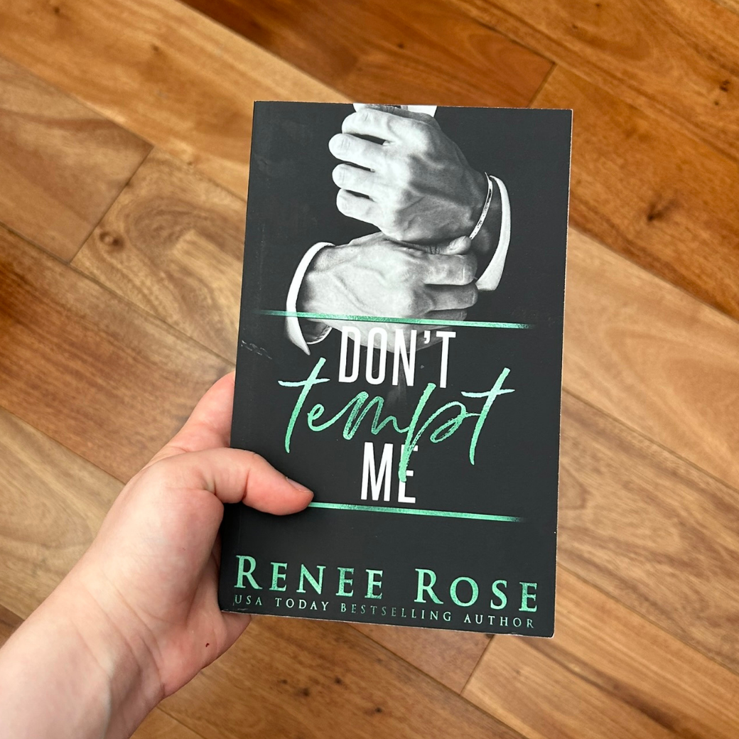 A white person's hand holding a paperback. The book cover has a black background and features a suited man's hands adjusting his bracelet. In white and green text is the title "Don't Tempt Me." On the bottom of the cover in green text is "Renee Rose -- USA Today Bestselling Author"