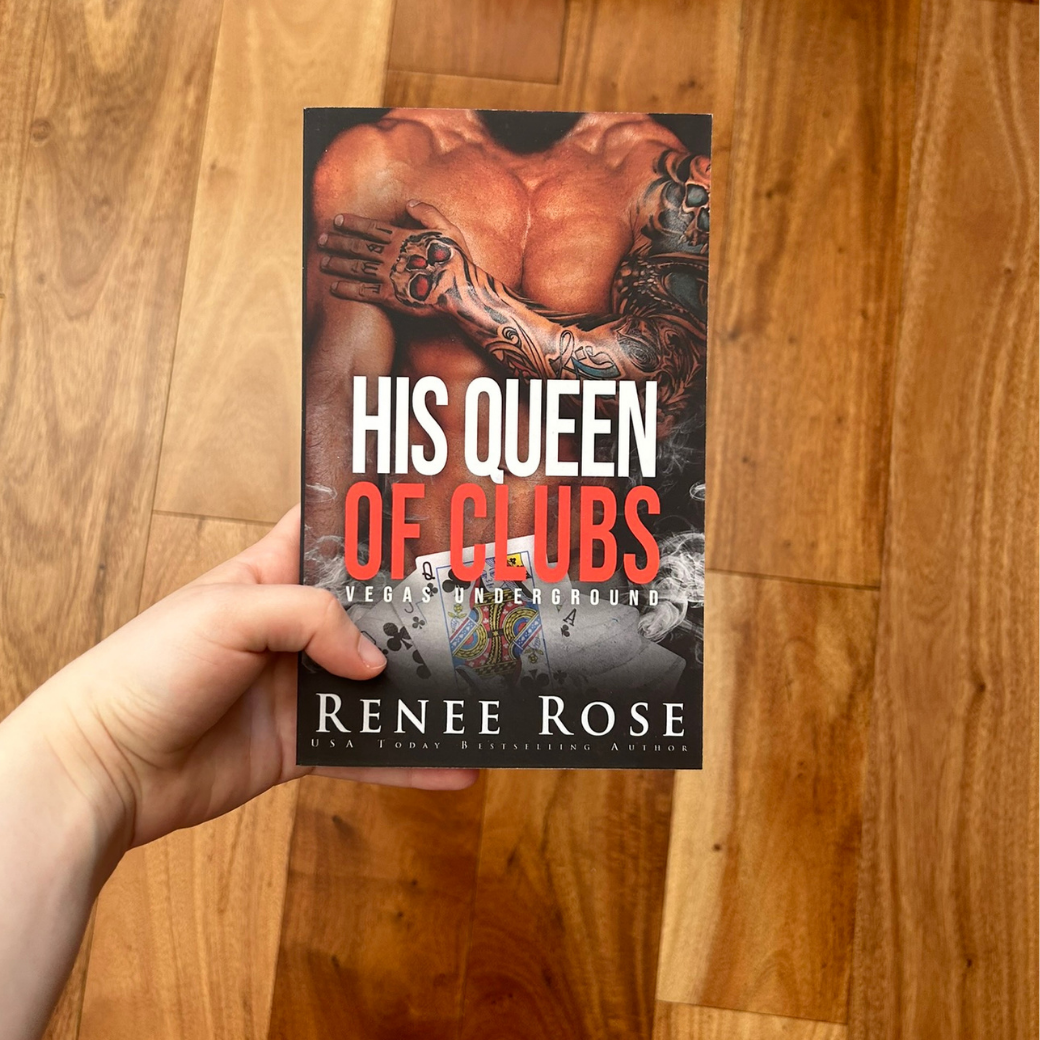 A white person's hand holding a paperback. The book cover features a buff, shirtless man who has an arm tattoo sleeve. Underneath him is some playing cards featuring a queen of clubs. In red and white text is the title "His Queen of Clubs Vegas Underground." On the bottom in white text is "Renee Rose -- USA Today Bestselling Author"