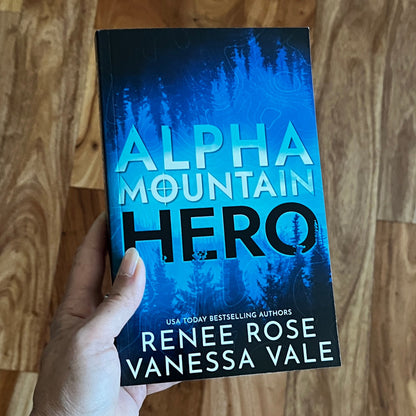 Book titled 'Alpha Mountain Hero' by Renee Rose and Vanessa Vale held by a hand on a wooden surface.