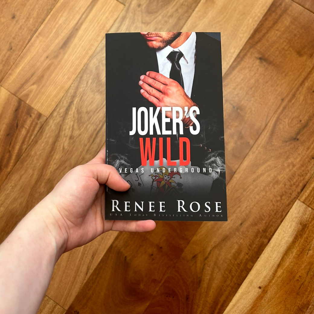 A white person's hand holding up a paperback. The cover features a man in a black suit and tie. He is adjusting his suit. Underneath him is a card deck with the joker on top. In white and red text is the title "Joker's Wild Vegas Underground." On the bottom in white is "Renee Rose -- USA Today Bestselling Author"