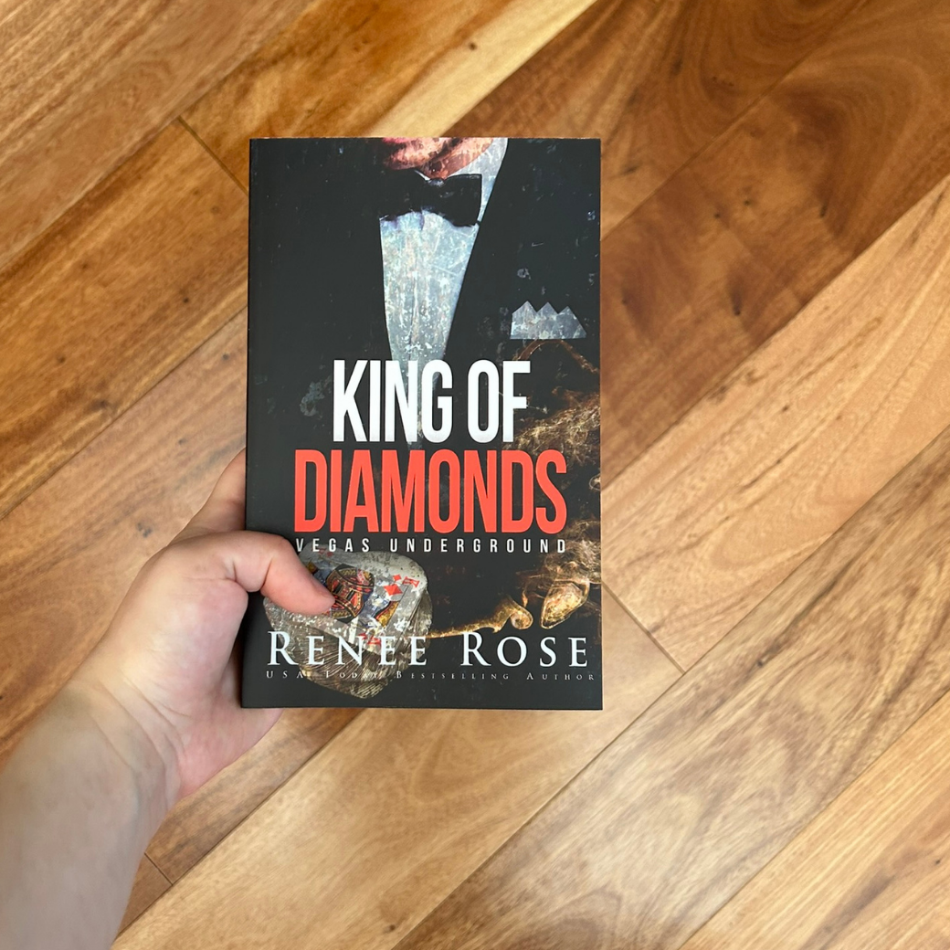 A white person's hand holding a paperback. The book cover features a man in a black and white suit and a black bow tie. Underneath him is a king of diamonds card. In white and red text is the title "King of Diamonds Vegas Underground." On the bottom in white is "Renee Rose -- USA Today Bestselling Author"