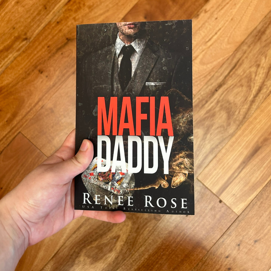 A white person's hand is holding up a paperback. The book cover features a man in a black and white suit. Underneath him is a deck of cards and fire. In red and white text is the title "Mafia Daddy." On the bottom of the cover in white text is "Renee Rose -- USA Today Bestselling Author"