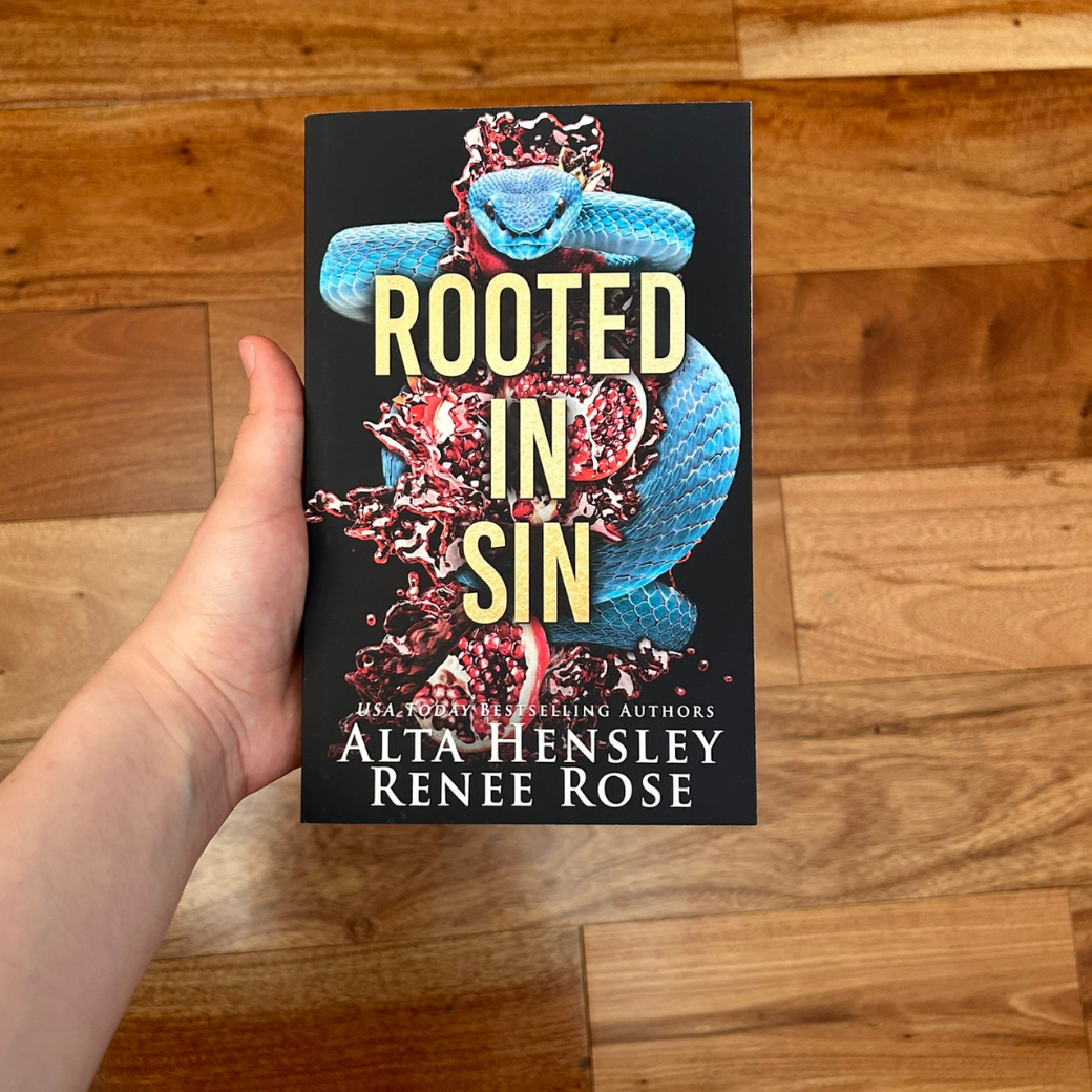 A white person's hand holding a paperback. The book cover's background is back. In stark contrast is a blue snake and pomegranate. On top of the design in gold text is "Rooted in Sin." On the bottom of the cover in white text "USA Today Bestselling Authors -- Alta Hensley Renee Rose" 