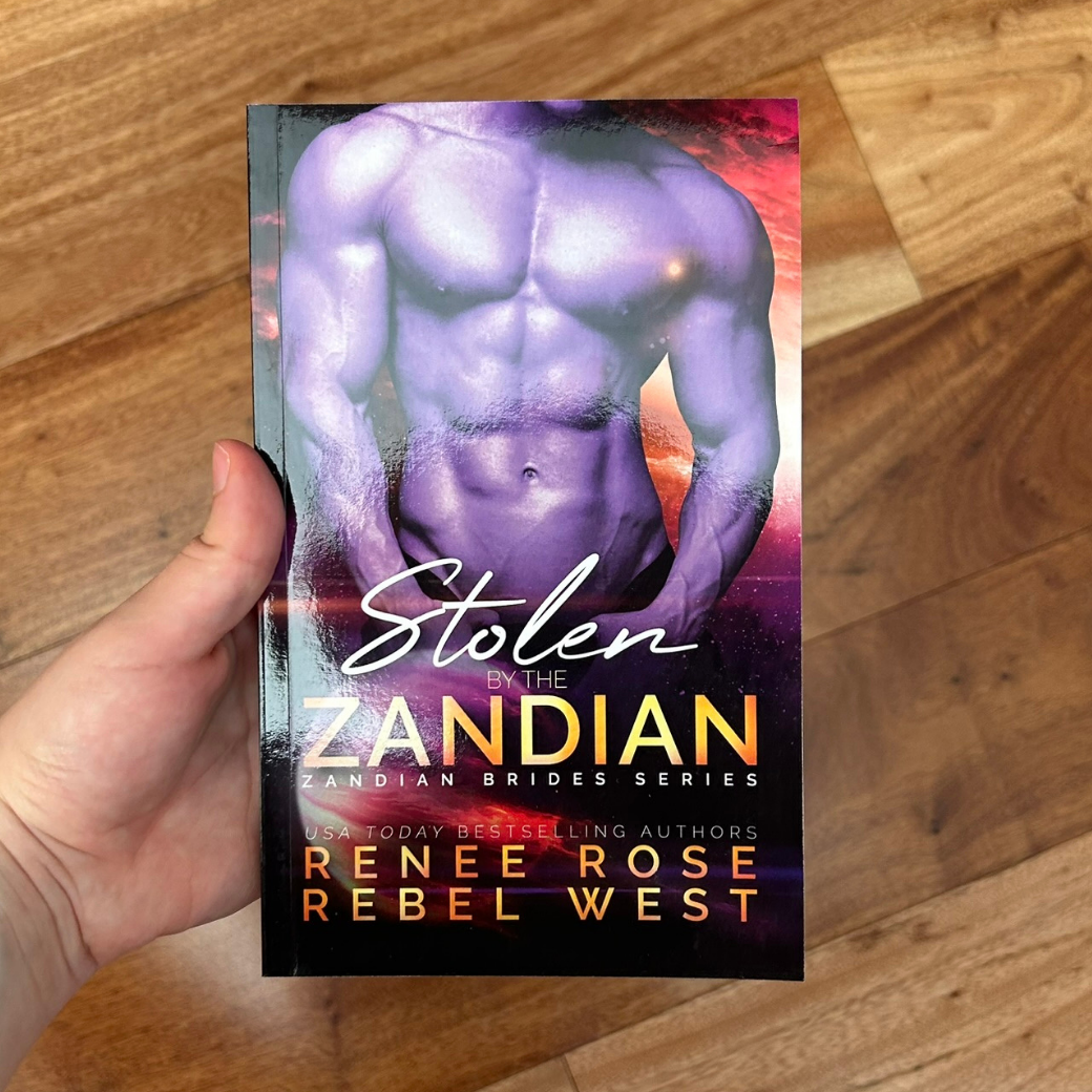 A white person's hand holding a paperback. The book cover features a buff, shirtless, white man in purple lighting. The background features a sunset and a planet. In white and gold text is the title "Stolen by the Zandian Zandian Brides Series." On the bottom of the cover in white and gold text is "USA Today Bestselling Authors --Renee Rose Rebel West"