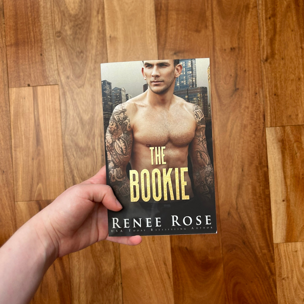 A white person's hand holding a paperback. The book cover features a white, buff, shirtless, man with arm tattoo sleeves. In gold text is the title "The Bookie." On the bottom of the cover in white text "Renee Rose -- USA Today Bestselling Author"