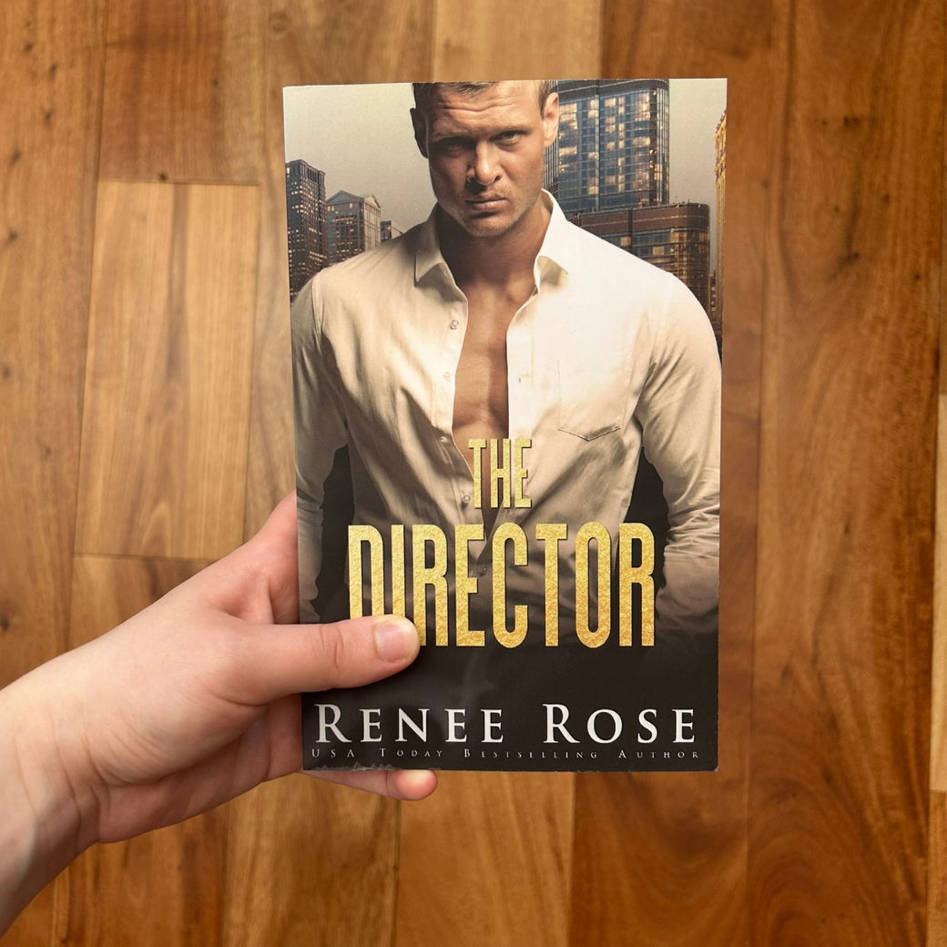 A white person's hand holding a paperback. The book cover features a buff white man looking out. He has a white unbuttoned shirt on. The background is of a city. In gold text is the title "The Director." On the bottom in white text is "Renee Rose -- USA Today Bestselling Author"