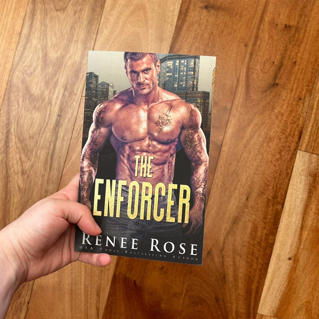 A white person's hand holding a paperback. The book cover features a buff, shirtless, tattooed man. In gold text is the title "The Enforcer." On the bottom of the cover in white text is "Renee Rose -- USA Today Bestselling Author"