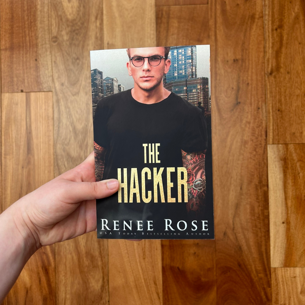 A white person's hand is holding a paperback. The book cover features a white man in a tight black shirt and wearing black glasses. He has arm tattoo sleeves. In gold text is the title "The Hacker." On the bottom in white text is "Renee Rose -- USA Today Bestselling Authors"