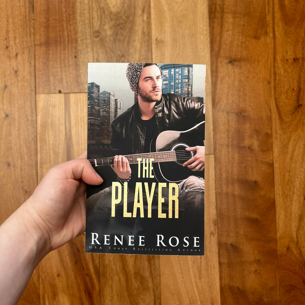 A white person's hand holding a paperback. The book cover features a man with scruff in a black shirt, black leather jacket, ripped jeans, beanie, and playing a black guitar. In gold text is the title "The Player." On the bottom of the cover in white text is "Renee Rose -- USA Today Bestselling Author"