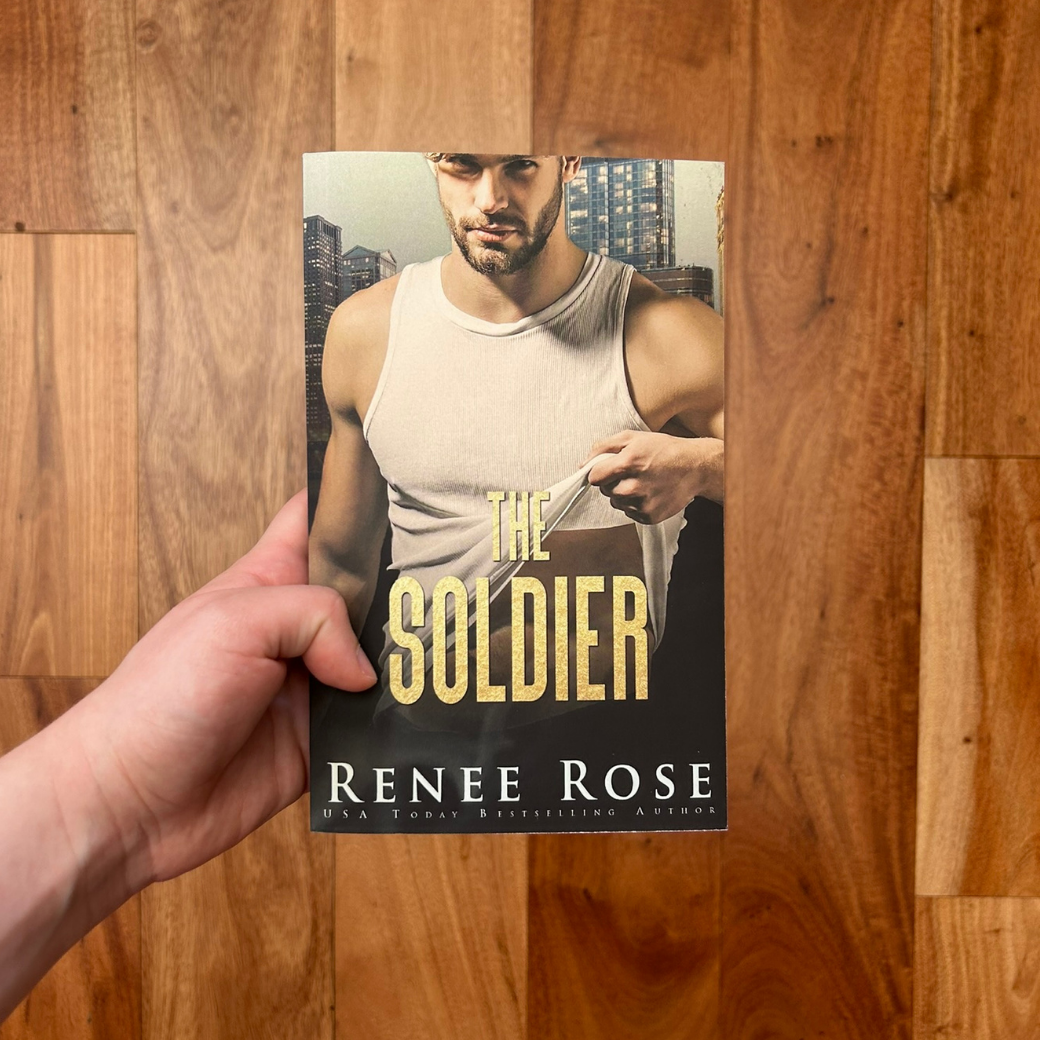 A white person's hand holding a paperback. The cover features a buff white man with black scruff. He is wearing a white tank top. The background is of a city. In gold text is the title "The Soldier." On the bottom in white text is "Renee Rose -- USA Today Bestselling Author"