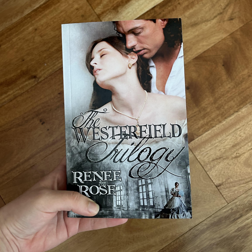 A white person's hand holding a paperback. The book cover features a white women in a white dress with a locket necklace and gold earrings. She is a brunette and is leaning her head against a man's chest. He is wearing a slightly unbuttoned white shirt and has long brown curly hair. In black text is the title "The Westerfield Trilogy." Underneath the title is an image of a women in a ball gown in a large room In white text is "Renee Rose"