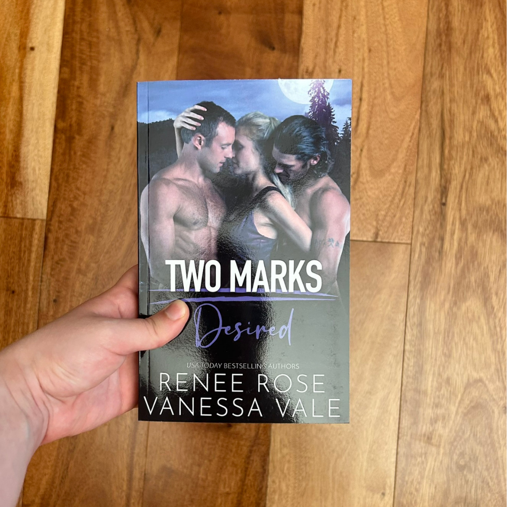 A white person's hand holding a paperback. Teh book cover features a blonde women wearing purple tank top. She is grabbing one buff, white, shirtless man's head. Behind her another man is grabbing her arm. In white and purple text is the title "Two Marks Desired." On the bottom in white text is "USA Today Bestselling Authors -- Renee Rose Vanessa Vale"