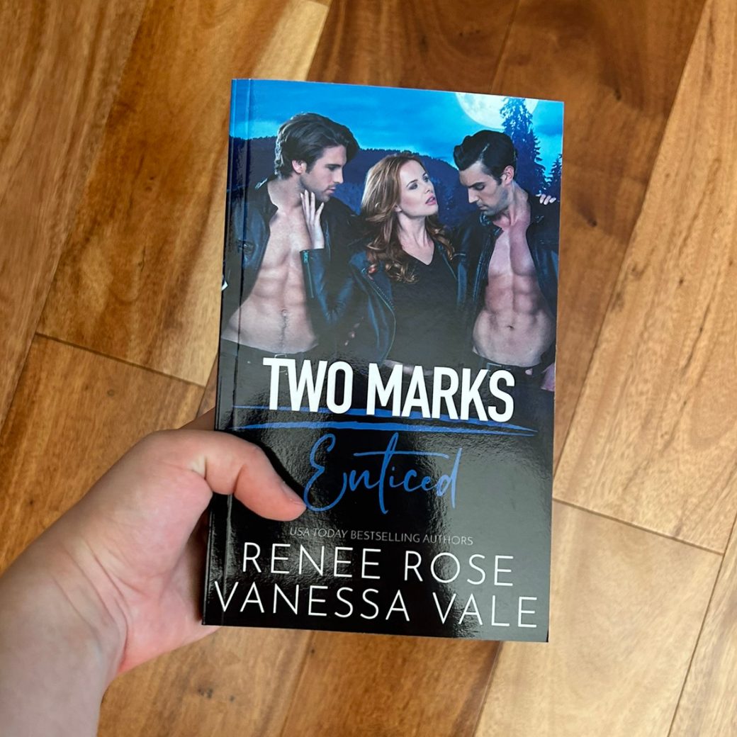 A white person's hand holding a paperback. The book cover features a red haired white women in a black shirt and black leather jacket. She is holding onto two white, buff, shirtless men who are wearing leather jackets and jeans. In white and blue text is the title "Two Marks Enticed." On the bottom of the cover in white text is "USA Today Bestselling Authors -- Renee Rose Vanessa Vale"