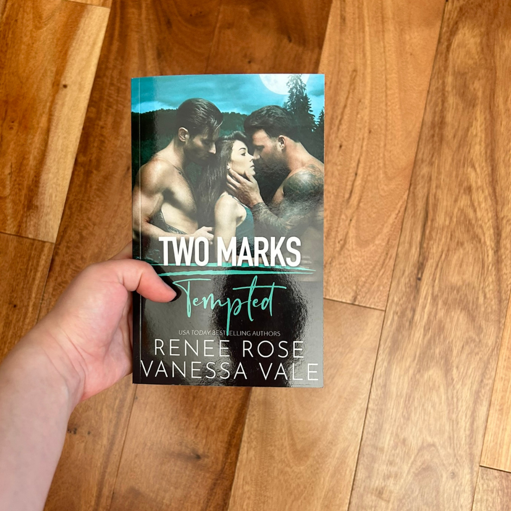 A white person's hand holding a paperback. The book cover features a brunette women wearing a grey tank top being grasped by two shirtless, buff, tattooed men. In the background is a full moon and a forest. In white and green text is the title "Two Marks Tempted." On the bottom of the cover in white text is "USA Today Bestselling Authors -- Renee Rose Vanessa Vale"