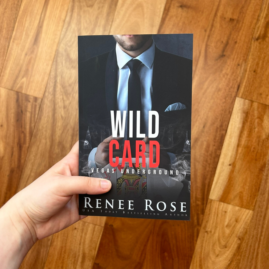 A white person's hand holding a paperback. The book cover features a white man in a black and white suit and tie. In white and red text is the title "Wild Card Vegas Underground." On the bottom of the cover in white text is "Renee Rose -- USA Today Bestselling Author"