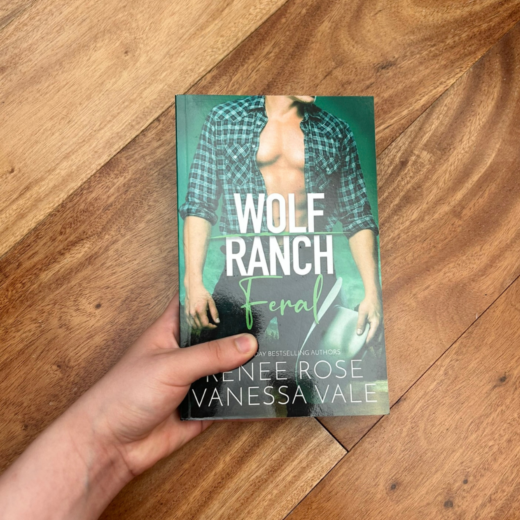 A white person's hand holding a paperback. The book cover's background is green. It features a buff white man wearing black jeans and an unbuttoned green flannel. He is holding a white cowboy hat in his hand. In white and green text is the title "Wolf Ranch Feral." On the bottom of the cover in white text is "USA Today Bestselling Authors -- Renee Rose Vanessa Vale"