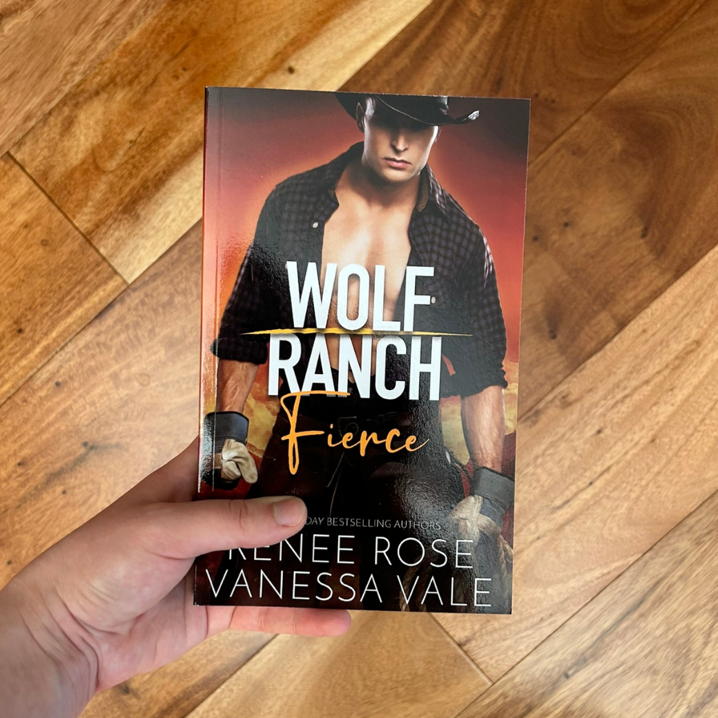 A white person's hand holding a paperback. The book cover's background is fire and orange. It features a white buff man in a black cowboy hat, black unbuttoned flannel, jeans, and worker gloves. In white and orange text is the title "Wolf Ranch Fierce." On the bottom of the cover in white text "USA Today Bestselling Authors -- Renee Rose Vanessa Vale"