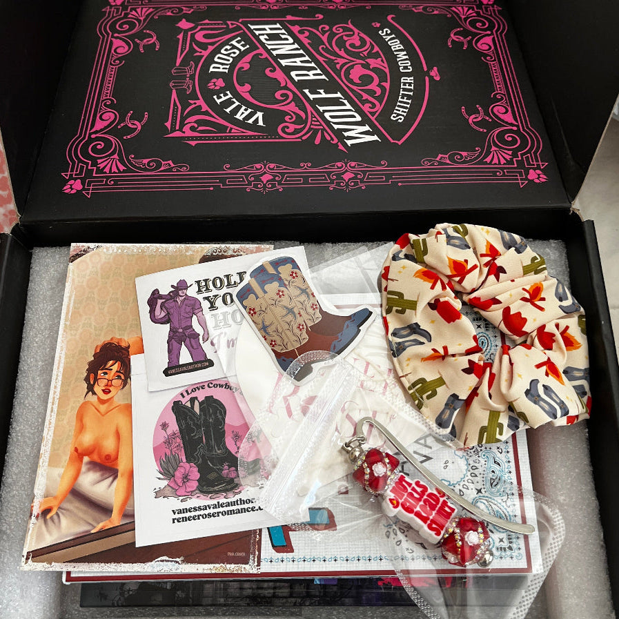 An open black gift box filled with assorted items, including a black lid printed with pink gothic text, printed NSFW character art card, a western scrunchie, 3 books, stickers, and a "hot Girls Read Smut" bookmark. 