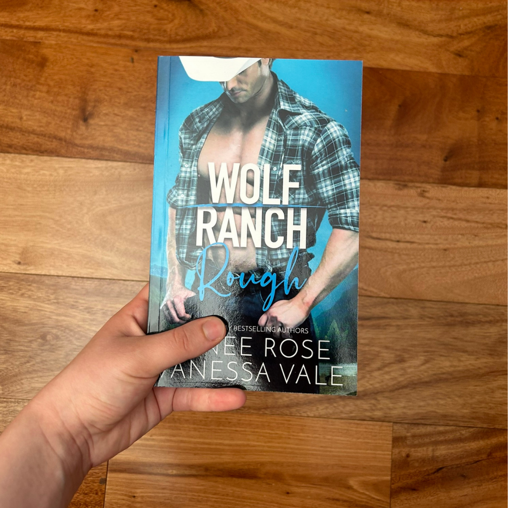 A white person's hand holding a paperback. The book cover's background is light blue. It features a buff white man wearing jeans, an unbuttoned flannel shirt, and a white cowboy hat. In white and blue text is the title "Wolf Ranch Rough." On the bottom of the cover in white text is "USA Bestselling Authors -- Renee Rose Vanessa Vale"