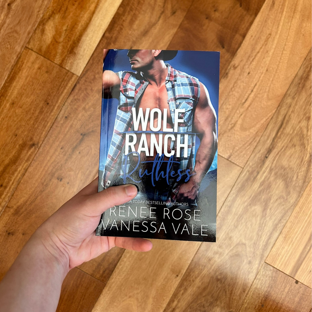 A white person's hand holding a paperback. The book cover's background is blue and mountains. It features a buff white man who is wearing a cowboy hat, unbuttoned blue flannel and jeans. In white and blue text is the title "Wolf Ranch Ruthless." On the bottom of the cover in white text is "USA Today Bestselling Authors -- Renee Rose Vanessa Vale"