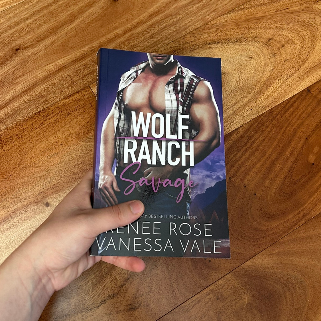 A white person's hand wearing a paperback. The book cover's background is purple. It features a buff, white man. He is wearing jeans and an unbuttoned white and red flannel. In white and purple text is the title "Wolf Ranch Savage." On the bottom of the cover in white text is "USA Today Bestselling Authors -- Renee Rose Vanessa Vale"