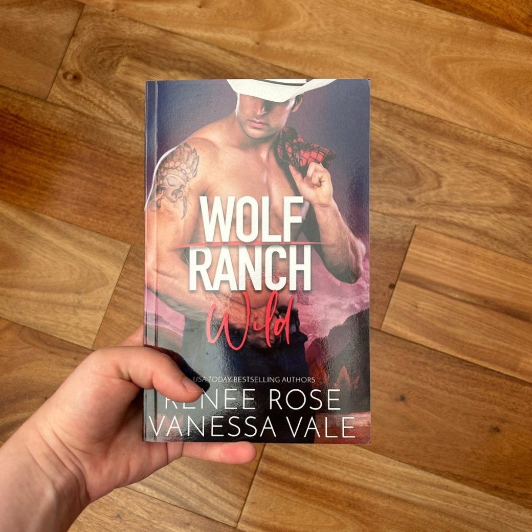 A white person's hand holding a paperback. The books cover's background is of a desert and pink/purple sky. It features a shirtless, buff, tattooed white man. He is wearing a white cowboy hat and is holding a red flannel over his shoulder. In white and red text is the title "Wolf Ranch Wild." On the bottom of the cover in white text "USA Today Bestselling Authors -- Renee Rose Vanessa Vale"